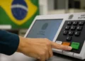 Mão de eleitor pressionando botão de confirmação em urna eletrônica durante votação no Brasil