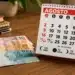 Calendário de agosto de 2026 ao lado de cédulas de real sobre uma mesa, ilustrando o calendário de pagamentos do Bolsa Família, INSS e Pé-de-Meia.