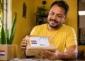 Homem segura encomenda com bandeira do Paraguai e etiqueta dos Correios dentro de casa.