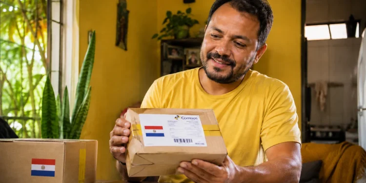 Homem segura encomenda com bandeira do Paraguai e etiqueta dos Correios dentro de casa.