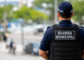 Guarda municipal fardado de costas durante patrulhamento, com o ambiente urbano desfocado ao fundo.