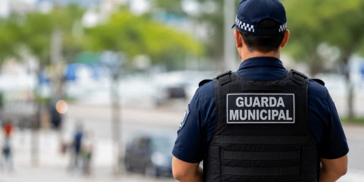 Guarda municipal fardado de costas durante patrulhamento, com o ambiente urbano desfocado ao fundo.