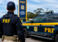 Policial Rodoviário Federal ao lado de viatura da PRF durante operação em rodovia.