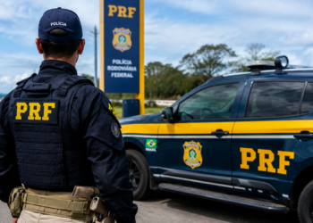 Policial Rodoviário Federal ao lado de viatura da PRF durante operação em rodovia.
