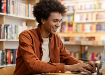 Estudante se prepara para concursos públicos usando notebook em biblioteca com livros ao fundo.