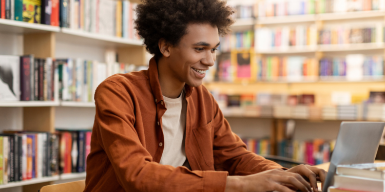 Estudante se prepara para concursos públicos usando notebook em biblioteca com livros ao fundo.