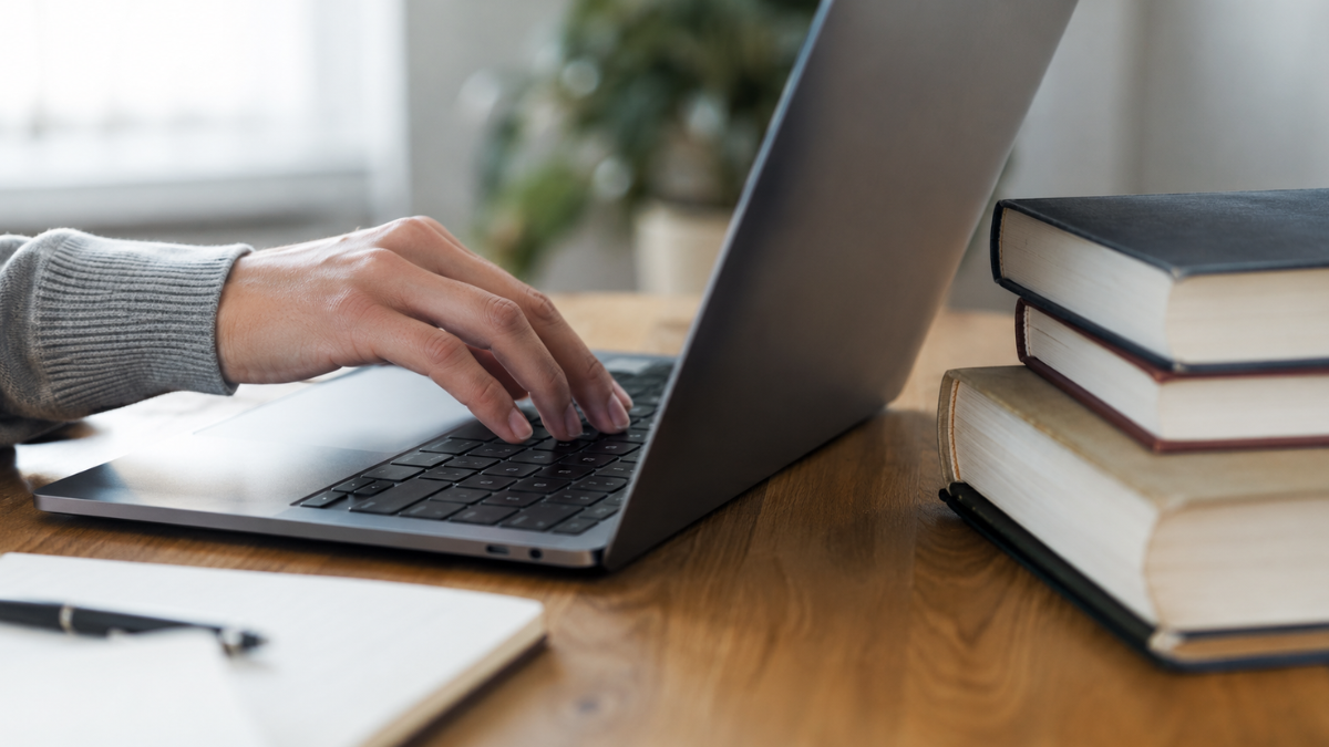 Mão digitando em notebook ao lado de livros, em referência às oportunidades em concursos de Câmaras Municipais.