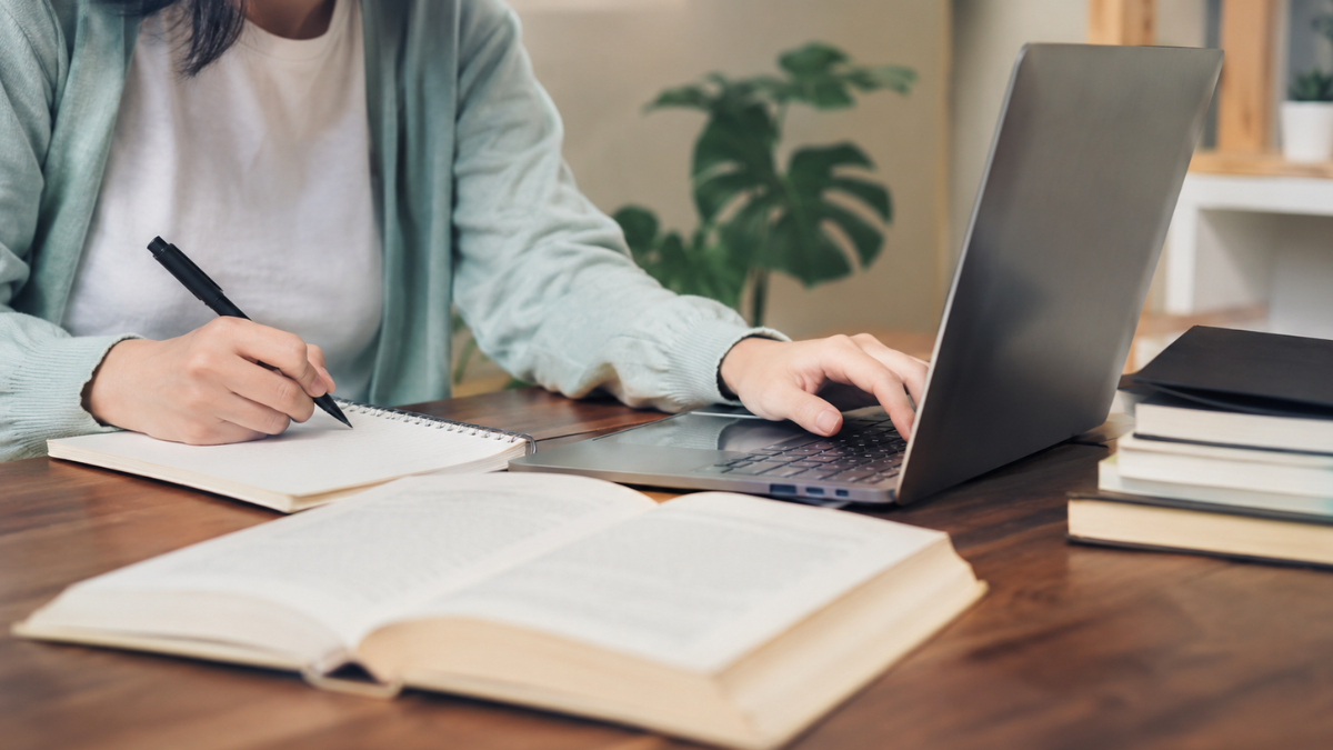 Pessoa estudando no notebook com livros e caderno de anotações para concurso público.