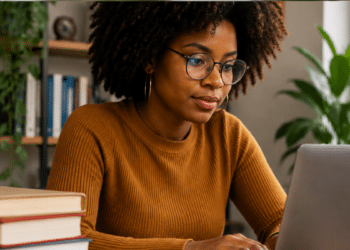 Mulher negra com cabelo afro e óculos estudando para concursos públicos usando notebook e livros em ambiente de estudo.