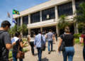 Pessoas se dirigem a uma Câmara Municipal com documentos e mochilas em busca de informações sobre concursos públicos.