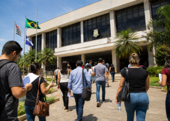 Pessoas se dirigem a uma Câmara Municipal com documentos e mochilas em busca de informações sobre concursos públicos.