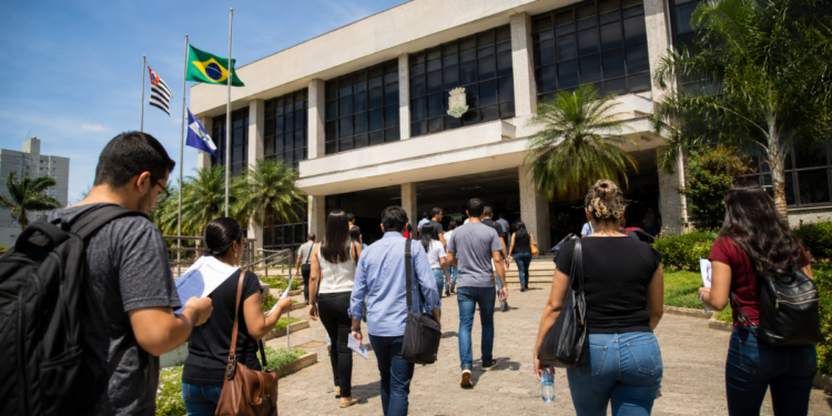 Pessoas se dirigem a uma Câmara Municipal com documentos e mochilas em busca de informações sobre concursos públicos.