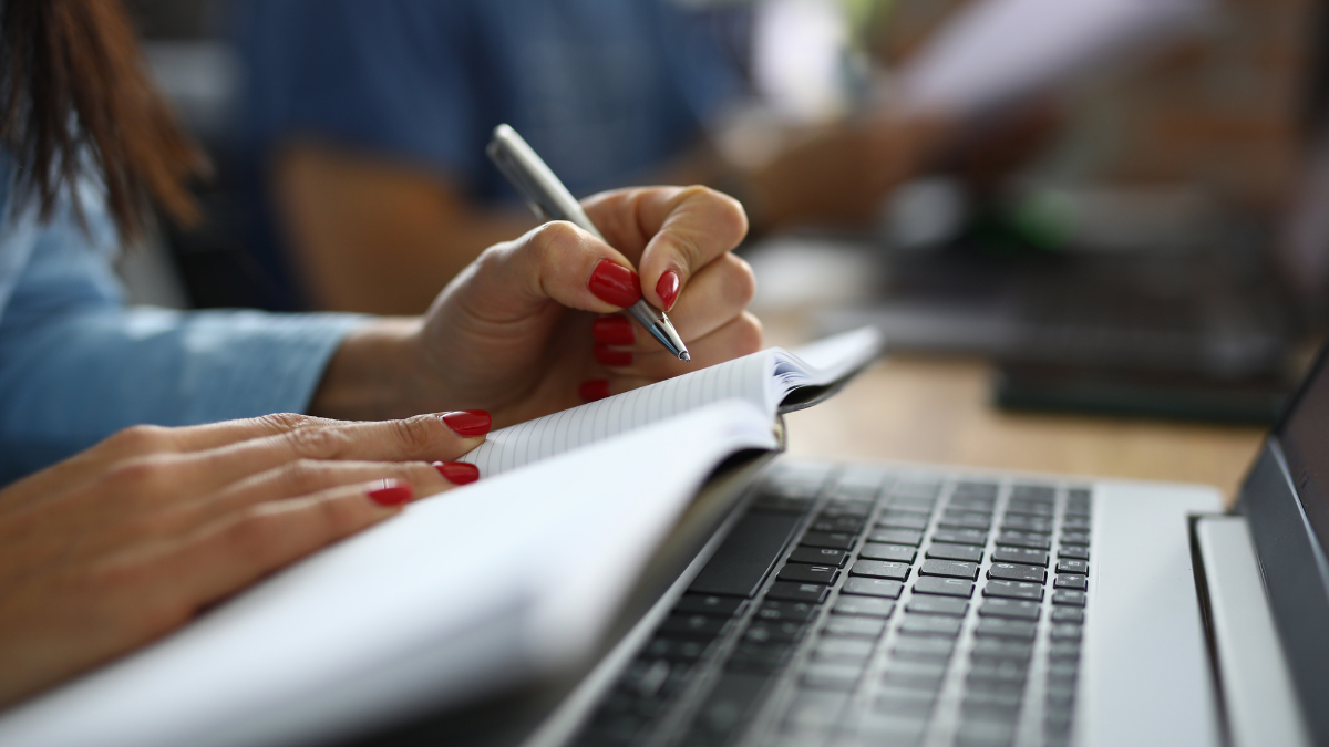 Pessoa faz anotações em caderno ao lado de notebook durante preparação para concurso público.