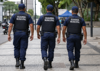 Três guardas municipais fardados caminham durante patrulhamento em área urbana.