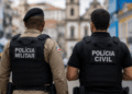 Policiais militar e civil da Bahia vistos de costas em uma área urbana.