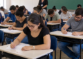 Pessoas realizando prova de concurso público em sala de aula com candidatos concentrados durante avaliação.