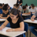 Pessoas realizando prova de concurso público em sala de aula com candidatos concentrados durante avaliação.