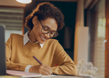 Mulher estudando em casa com caderno e notebook para concursos públicos.