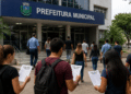 Pessoas caminhando em direção à prefeitura municipal com documentos de concurso público em mãos.