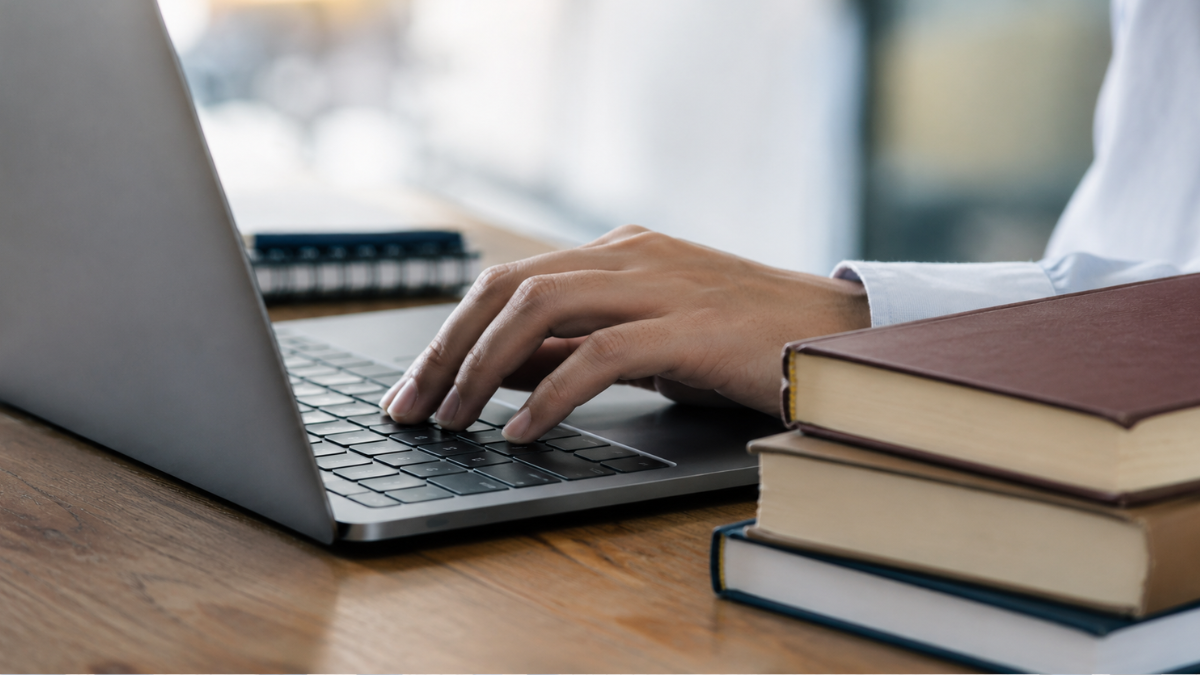 Mão digitando em notebook ao lado de livros representa preparação para concursos públicos em São Paulo.