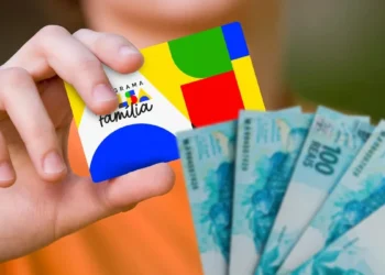 Mão segura cartão do Bolsa Família ao lado de cédulas de real, em referência à continuidade dos pagamentos do programa.
