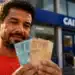 Trabalhador informal segura notas de real em frente a uma agência bancária, representando o acesso ao crédito pelo Desenrola Adimplentes.