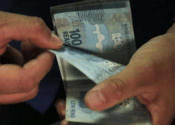 Imagem ilustrativa de mãos contando notas de cem reais, cena ligada ao montante de valores esquecidos disponível para resgate no sistema financeiro.