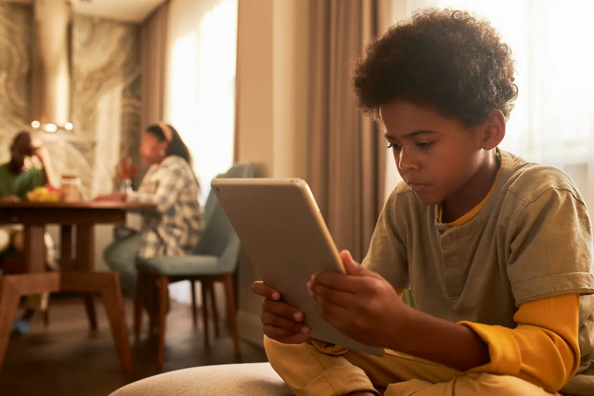 Criança utiliza tablet em casa sob supervisão de adultos ao fundo, representando proteção e segurança no ambiente digital.