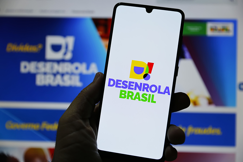 Imagem ilustrativa de mão segurando celular com a marca do Desenrola Brasil na tela, diante do site do programa exibido no monitor, em referência ao público habilitado a aderir