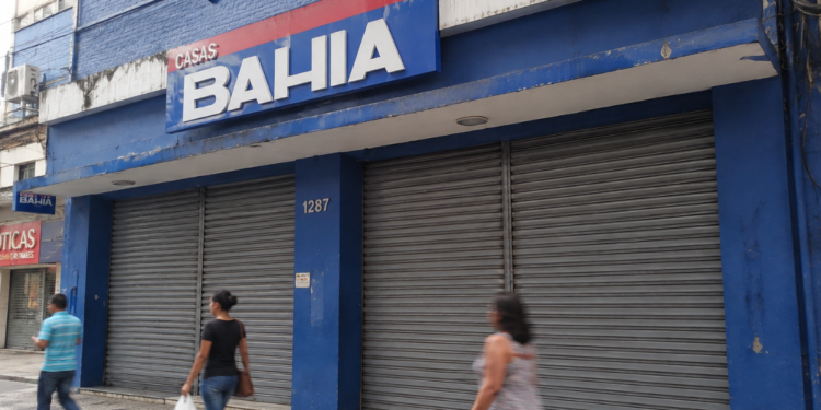 Fachada de loja da Casas Bahia com portas de ferro fechadas e pedestres passando pela calçada.