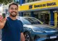 Motorista segura chave ao lado de carro elétrico BYD em frente a uma agência do Banco do Brasil.