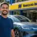 Motorista segura chave ao lado de carro elétrico BYD em frente a uma agência do Banco do Brasil.