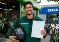 Jovem entregador segura capacete e contrato de financiamento diante de motos e bicicletas elétricas, com agência da Caixa ao fundo.