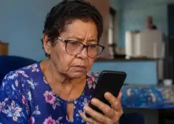 Idosa observa celular após receber mensagem suspeita sobre corte de benefício do INSS por falta de Prova de Vida.