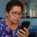 Idosa observa celular após receber mensagem suspeita sobre corte de benefício do INSS por falta de Prova de Vida.