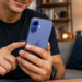 Homem sorrindo interage com iPhone azul enquanto está sentado em sofá, com luminária ao fundo.