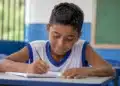 Estudante do ensino fundamental escreve em sala de aula durante atividade escolar sobre o desempenho da educação básica em tempo integral.