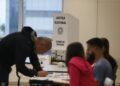 Seção eleitoral em funcionamento, com cabina de votação da Justiça Eleitoral ao centro, mesários sentados à mesa e eleitor assinando a lista de presença.