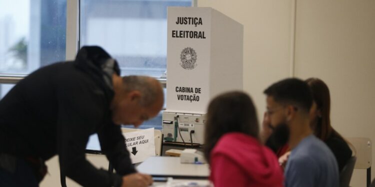 Seção eleitoral em funcionamento, com cabina de votação da Justiça Eleitoral ao centro, mesários sentados à mesa e eleitor assinando a lista de presença.