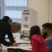 Seção eleitoral em funcionamento, com cabina de votação da Justiça Eleitoral ao centro, mesários sentados à mesa e eleitor assinando a lista de presença.
