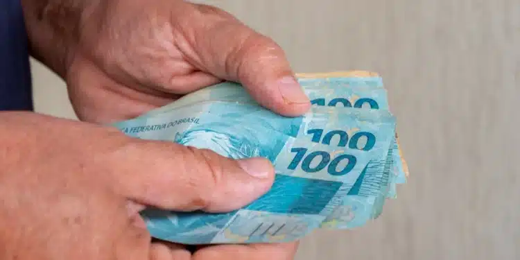 Mãos segurando cédulas de R$ 100 em referência à nova previsão do salário mínimo de 2027.