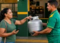Mulher recebe botijão de gás em revendedora com celular na mão, representando a consulta e o recebimento do benefício Gás do Povo em setembro.