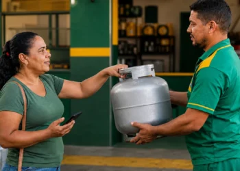 Mulher recebe botijão de gás em revendedora com celular na mão, representando a consulta e o recebimento do benefício Gás do Povo em setembro.