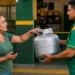 Mulher recebe botijão de gás em revendedora com celular na mão, representando a consulta e o recebimento do benefício Gás do Povo em setembro.