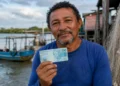 Pescador artesanal segura nota de 100 reais próximo à embarcação, representando o pagamento do segundo lote do seguro-desemprego do INSS.