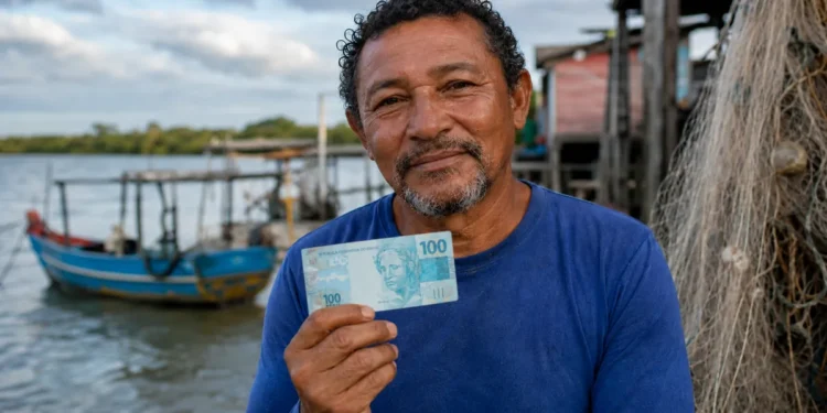 Pescador artesanal segura nota de 100 reais próximo à embarcação, representando o pagamento do segundo lote do seguro-desemprego do INSS.