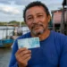 Pescador artesanal segura nota de 100 reais próximo à embarcação, representando o pagamento do segundo lote do seguro-desemprego do INSS.