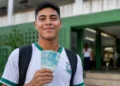 Estudante da EJA segura notas de R$ 100 em frente a uma escola, representando o pagamento do Pé-de-Meia.