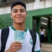 Estudante da EJA segura notas de R$ 100 em frente a uma escola, representando o pagamento do Pé-de-Meia.
