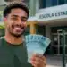 Estudante da EJA sorrindo em frente a uma escola e segurando cédulas, em referência ao pagamento de R$ 1.000 do Pé-de-Meia.